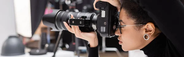 Vista lateral del fotógrafo afroamericano en anteojos trabajando con cámara digital en estudio fotográfico, pancarta - foto de stock