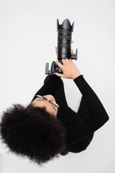 Вид сверху на африканского американского фотографа в черной одежде, фотографирующего на цифровую камеру, изолированную на сером — стоковое фото