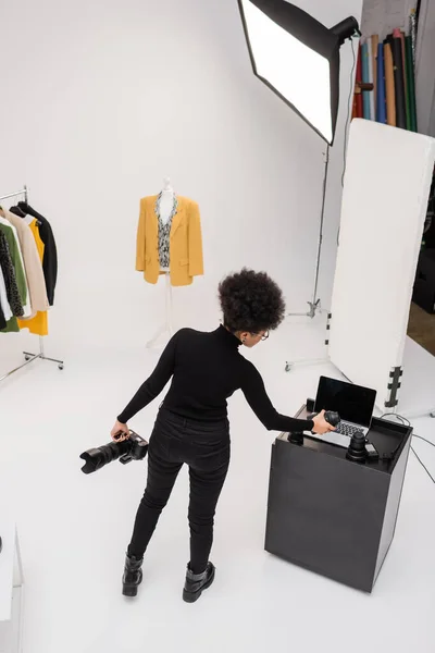 Vue grand angle du photographe afro-américain avec appareil photo numérique tenant objectif photo près d'un ordinateur portable dans un studio photo moderne — Photo de stock