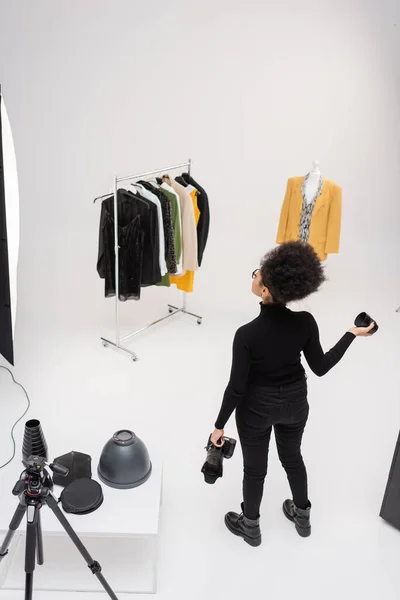 Vista ad alto angolo del creatore di contenuti afro-americani con fotocamera digitale e obiettivo fotografico guardando collezione di vestiti alla moda in studio fotografico — Foto stock