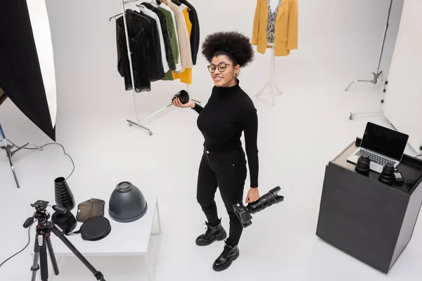 Vue grand angle du photographe afro-américain heureux tenant appareil photo numérique et objectif photo près de vêtements élégants dans le studio photo — Photo de stock