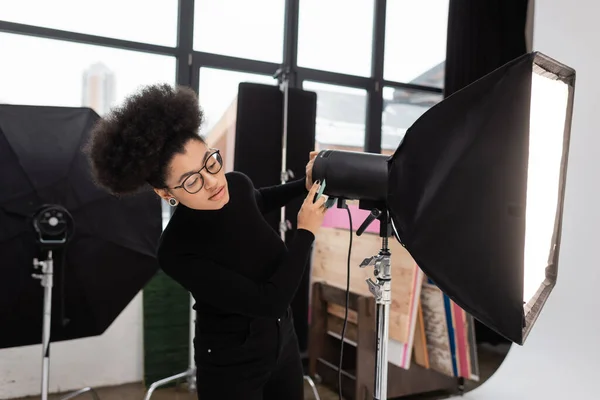 Producteur de contenu afro-américain dans les lunettes de travail avec réflecteur softbox dans le studio photo — Photo de stock