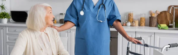Infermiera multirazziale in uniforme blu con stetoscopio in piedi vicino a donna anziana e walker, striscione — Foto stock