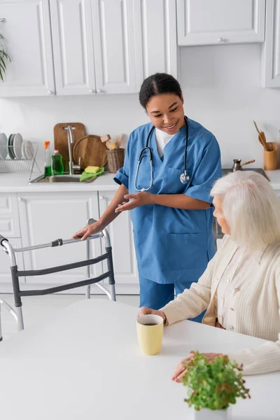 Fröhliche Krankenschwester in blauer Uniform, die mit der Hand auf Rollator zeigt, während sie neben einer Seniorin mit grauen Haaren steht — Stockfoto