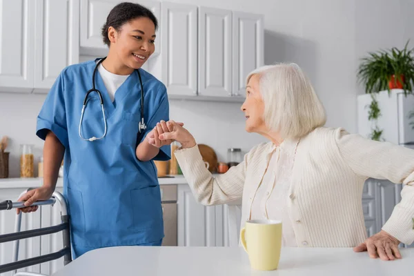 Glückliche multirassische Krankenschwester in blauer Uniform, die Hand einer Seniorin mit grauen Haaren hält, während sie neben einem Rollator steht — Stockfoto