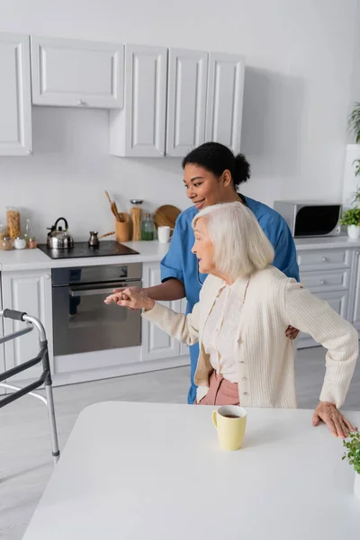 Щасливий багаторасовий доглядач у блакитній формі тримає руку старшої жінки з сірим волоссям, стоячи біля ходунки — стокове фото