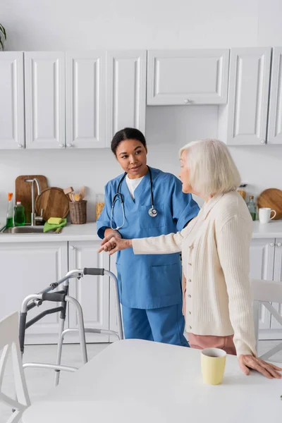 Brünette, multiethnische Pflegerin in blauer Uniform unterstützt Seniorin mit grauen Haaren, während sie neben Rollator steht — Stockfoto