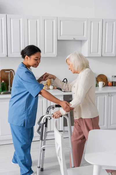 Брюнетка многорасовая медсестра в синей форме поддерживает пожилую женщину с седыми волосами, стоящую рядом с ходунком — стоковое фото