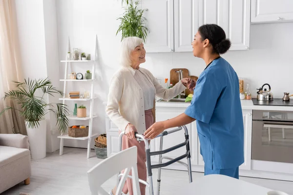 Брюнетка многорасовая медсестра в синей форме поддерживает счастливую пожилую женщину с седыми волосами, стоящую рядом с ходунком — стоковое фото