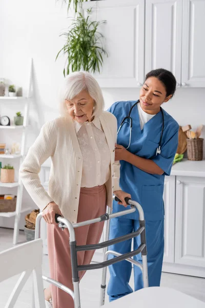 Brunette infirmière multiraciale en uniforme bleu soutenant la femme âgée avec les cheveux gris marchant avec l'aide de marcheur — Photo de stock