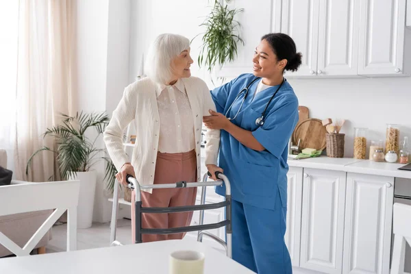 Heureuse infirmière multiraciale en uniforme bleu soutenant la femme âgée avec les cheveux gris marchant avec l'aide de marcheur — Photo de stock
