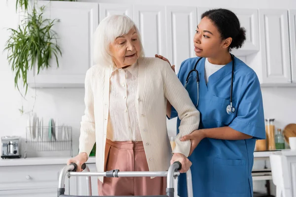 Brunette multiracial nurse in blue uniform supporting senior woman with grey hair walking with help of walker at home — Stock Photo
