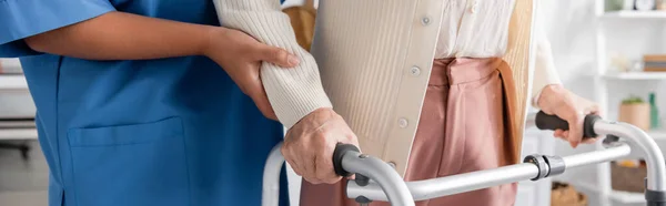 Обрезанный вид многорасовой медсестры в синей форме, поддерживающей пожилую женщину, идущую с помощью ходунков, баннера — стоковое фото