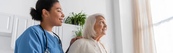 Alegre enfermera multirracial en uniforme azul mirando feliz mujer mayor con el pelo gris en casa, pancarta - foto de stock