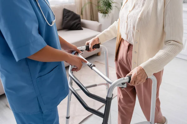 Partial view of multiracial nurse in blue uniform supporting senior woman walking with help of walker at home — Stock Photo