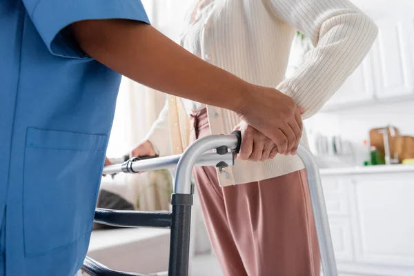Cropped view of multiracial caregiver in uniform supporting senior woman walking with walker at home — Stock Photo