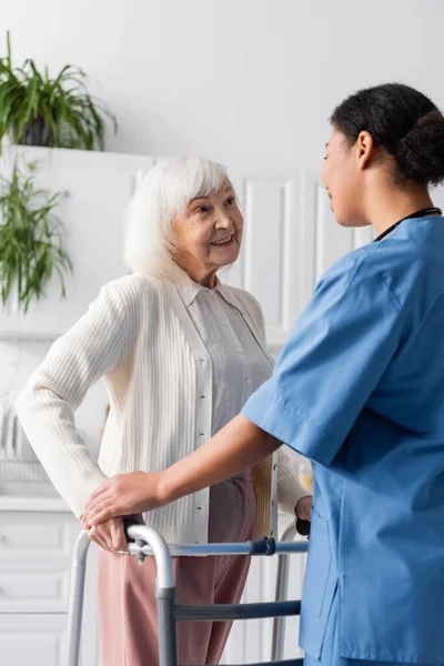 Счастливая пожилая женщина с седыми волосами ходит с рамкой ходунка и глядя на брюнетку многорасовую медсестру — стоковое фото