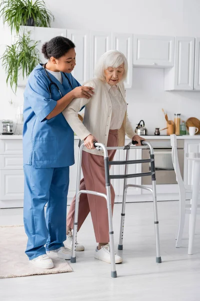 Повна довжина багаторасової медсестри в уніформі підтримуючої пенсіонерки з сірим волоссям, що йде з прогулянковою рамою вдома — стокове фото