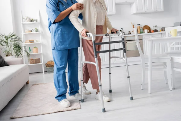 Recortado vista de cuidador multirracial en uniforme de apoyo a la mujer mayor caminando con andador en apartamento moderno - foto de stock