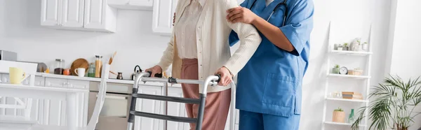 Обрізаний вид на багаторасового доглядальника в уніформі, що підтримує старшу жінку, що ходить з ходунками в сучасній квартирі, банер — стокове фото
