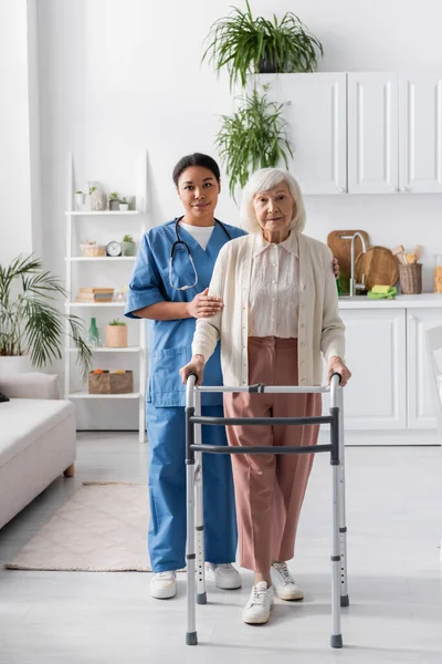 Longitud completa de la enfermera multirracial que apoya a la mujer jubilada con el pelo gris que camina con el marco que camina en casa - foto de stock