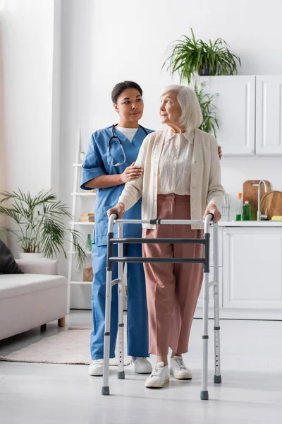 Intera lunghezza dell'infermiera multirazziale che sostiene la donna anziana con i capelli grigi che cammina con la struttura ambulante a casa — Foto stock