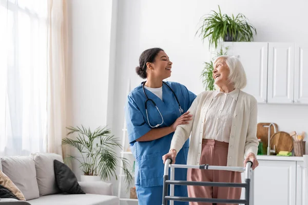 Happy multiracial nurse in uniform laughing with retired woman while standing near walking frame at home — Stock Photo