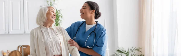 Happy multiracial nurse in uniform laughing with retired woman at home, banner — Stock Photo