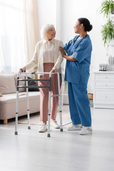 Full length of retired woman with grey hair walking with walking frame and looking at multiracial nurse in uniform — Stock Photo