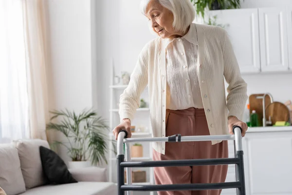 Mujer mayor con pelo gris caminando con ayuda de andador en apartamento moderno - foto de stock