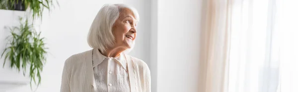 Mujer mayor feliz con el pelo gris mirando hacia otro lado mientras sonríe en casa, pancarta - foto de stock
