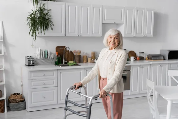 Положительная пожилая женщина с седыми волосами ходьба с помощью ходунков в современной квартире — стоковое фото