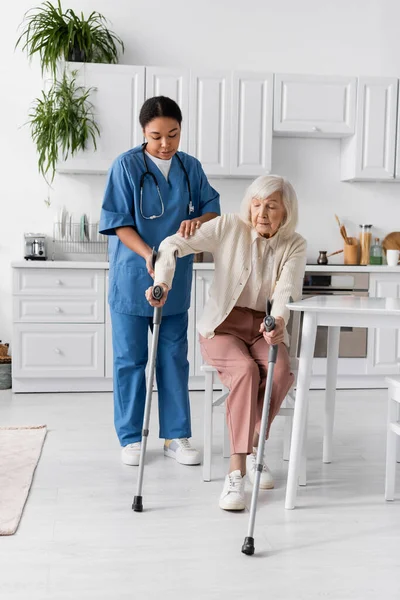 Повна довжина багаторасової медсестри в уніформі допомагає старшій жінці, використовуючи милиці, щоб встати — стокове фото
