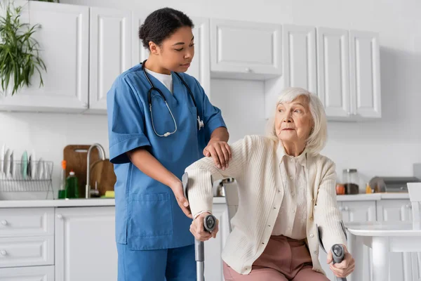 Багаторасовий медсестра в уніформі допомагає старшій жінці, використовуючи милиці, щоб встати в сучасній квартирі — стокове фото