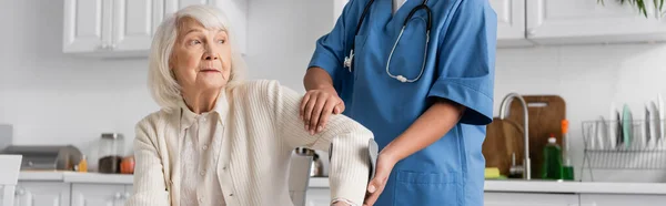 Infermiera in uniforme aiutare la donna anziana con stampelle per alzarsi in piedi in appartamento moderno, banner — Foto stock