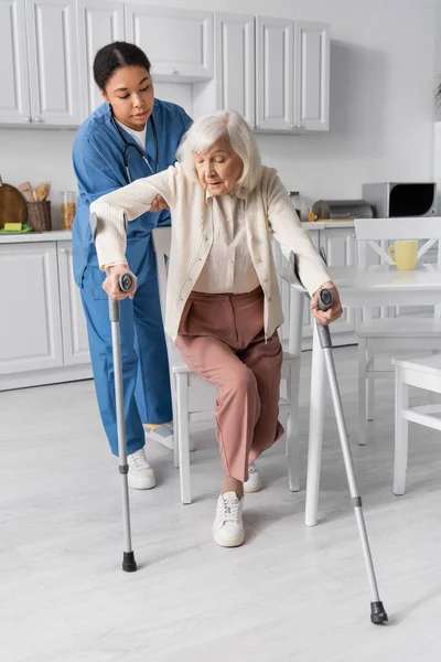 Longitud completa de cuidador multirracial en uniforme ayudar a las mujeres mayores que usan muletas para ponerse de pie en casa - foto de stock