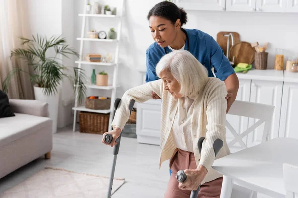 Brünette multiethnische Pflegerin in Uniform hilft Seniorin mit Krücken, zu Hause zu gehen — Stockfoto