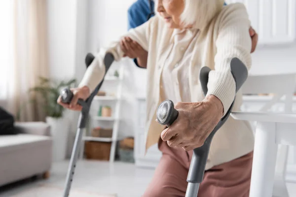 Visão cortada de cuidador multirracial em uniforme ajudando a mulher idosa usando muletas para andar em casa — Fotografia de Stock