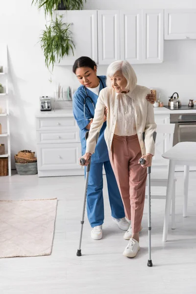 Comprimento total de cuidador multirracial em uniforme azul ajudando a mulher idosa usando muletas para andar em casa — Fotografia de Stock