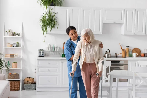 Brunette multiracial soignant en uniforme bleu aider la femme retraitée en utilisant des béquilles pour marcher dans l'appartement — Photo de stock