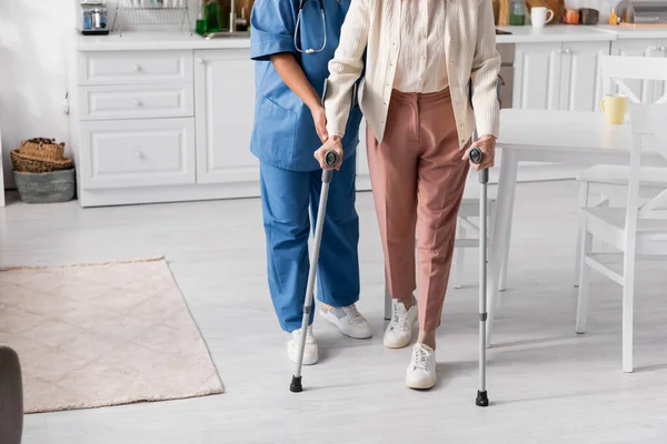 Обрезанный вид пожилой женщины с помощью костылей во время прогулки рядом с медсестрой дома — стоковое фото
