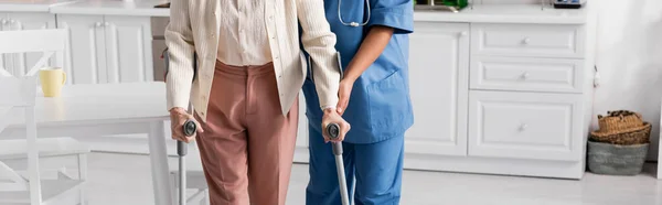 Cropped view of senior woman using crutches while walking near nurse at home, banner — Stock Photo