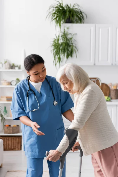 Retired woman with grey hair using crutches while walking near multiracial nurse at home — Stock Photo