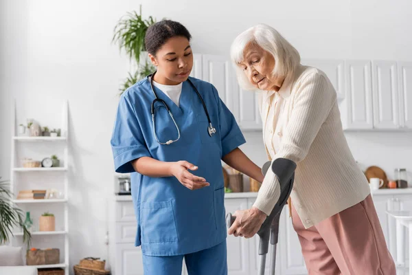 Seniorin mit grauen Haaren mit Krücken beim Gehen in der Nähe einer Krankenschwester — Stockfoto