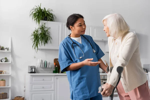 Retired woman with grey hair using crutches and talking with multiracial nurse at home — Stock Photo