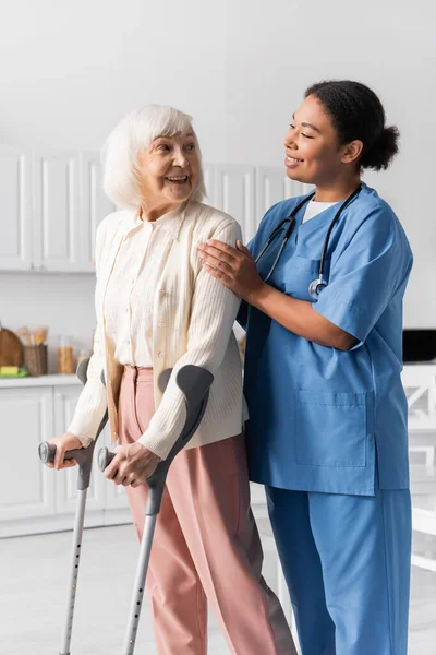 Cheerful retired woman with grey hair using crutches while walking near multiracial nurse at home — Stock Photo