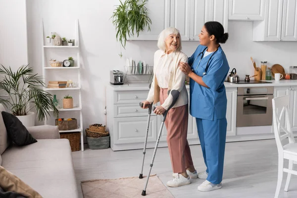 Longitud completa de mujer mayor alegre con pelo gris usando muletas mientras camina cerca de enfermera multirracial en casa - foto de stock
