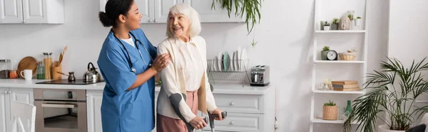 Alegre anciana con pelo gris usando muletas y caminando cerca de la enfermera multirracial en casa, pancarta - foto de stock