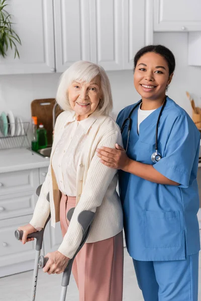 Mujer jubilada con pelo gris usando muletas y sonriendo mientras camina cerca de la enfermera multirracial feliz en casa - foto de stock