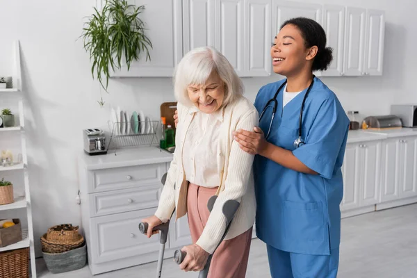 Mujer jubilada positiva con pelo gris usando muletas mientras camina cerca de la enfermera multirracial en casa - foto de stock